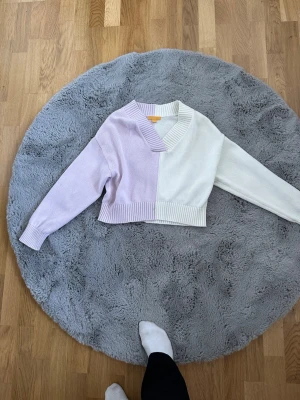 Ljuslila och vit cropped stickad tröja - Trendig stickad tröja i blockfärger, ljuslila och vit. Croppad modell med bred ribbstickad mudd i nederkant och ärmslut. V-ringad halsringning och lång ärm. Perfekt för en cool och avslappnad look.