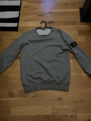 Grå sweatshirt från Stone Island - Säljer en stilren grå sweatshirt från Stone Island med den klassiska loggan på vänster ärm. Tröjan har rund halsringning, ribbade muddar och är tillverkad i mjukt bomullsmaterial. Perfekt för en avslappnad och trendig look.