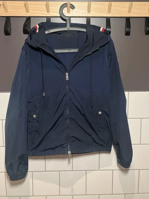 Marinblå vindjacka från Moncler - Snygg marinblå vindjacka från Moncler med huva och dragkedja framtill. Jackan har vita och röda detaljer på huvan, två fickor med tryckknappar och elastiska muddar vid ärmslut och midja. Pris kan diskuteras vid snabb affär