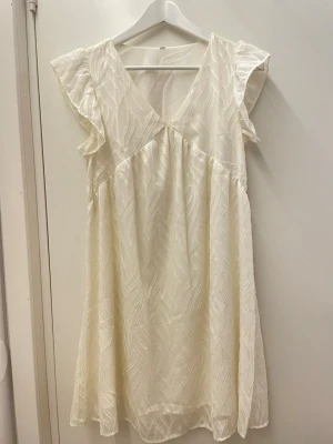 Vit kortklänning med volangärmar - Söt vit kortklänning med v-ringning och volangärmar. Klänningen har ett subtilt mönster i tyget och är tillverkad i ett lätt, luftigt material som passar perfekt till varmare dagar.