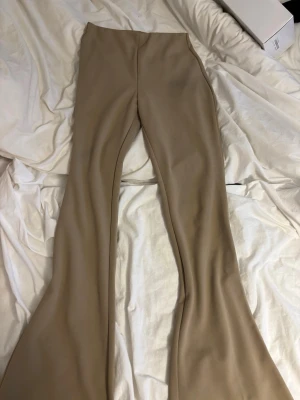 Beiga utsvängda byxor - Säljer ett par snygga beiga byxor med utsvängda ben. Byxorna har en hög midja och är tillverkade i ett stretchigt material som sitter skönt. Perfekta för dig som gillar trendiga och stilrena plagg med en retrovibe.