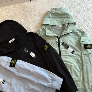 Stone Island vindjacka med huva - Snygg vindjacka från Stone Island med ikonisk kompasspatch på ärmen. Finns i svart, ljusgrått och ljusgrönt. Jackan har dragkedja framtill, huva och praktiska fickor. Tillverkad i lätt, vindtätt material som passar perfekt för blåsiga dagar.