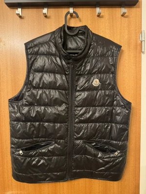 Svart dunväst från Moncler - Snygg svart dunväst från Moncler med quiltad design och hög krage. Västen har två dragkedjeförsedda fickor framtill och klassisk Moncler-logga på bröstet. Perfekt för lager-på-lager och ger en stilren look.