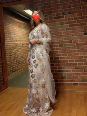 Kurdisk klänning, Vit långklänning med blommigt mönster - Elegant vit långklänning med blommigt mönster i guld och silver. Klänningen har transparenta ärmar och är tillverkad i ett glittrigt, tunt material som ger en lyxig känsla. Perfekt för speciella tillfällen och fester.