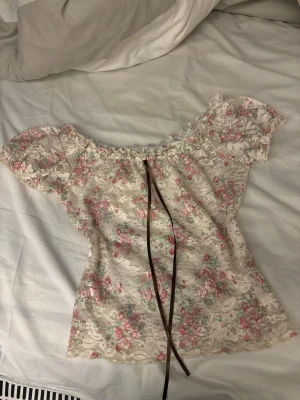 Blommig spetsblus med snörning - Supersöt blommig blus i vit spets med rosa och gröna detaljer. Offshoulder-modell med korta puffärmar och snörning framtill. Perfekt för dig som gillar romantisk och feminin stil.