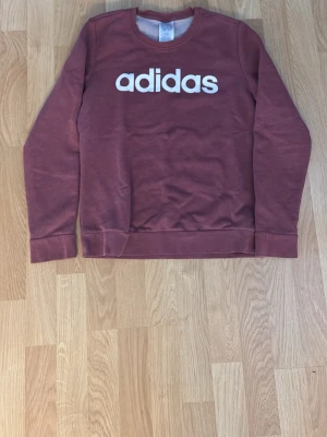 Vinröd stickad tröja från Adidas - Snygg vinröd stickad tröja från Adidas med vit logga framtill. Tröjan har rund halsringning, långa ärmar och ribbade muddar vid ärmslut och nederkant. Perfekt för en avslappnad och sportig stil.