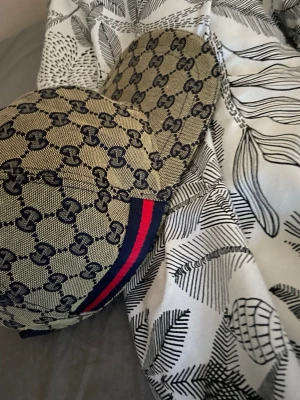 Gucci beige keps med GG-mönster - Säljer en beige keps från Gucci med klassiskt GG-monogram i mörkblått över hela kepsen. På sidan finns en ikonisk röd och marinblå rand. Kepsen har böjd skärm och är tillverkad i ett slitstarkt tygmaterial. Perfekt för dig som vill ha en lyxig och trendig accessoar.
