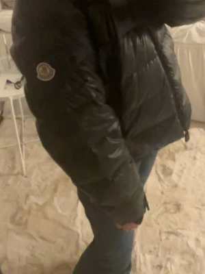 Svart pufferjacka från Moncler - Snygg svart pufferjacka från Moncler med glansig finish och klassisk Moncler-logga på ärmen. Jackan har huva, dragkedja framtill och två sidofickor med dragkedja. Perfekt för kalla vinterdagar och riktigt trendig look. köptes här på plick. vet ej om den är äkta! den har lite skador men om nån har någon fråga elelr om priset är det bara att höra av sig!