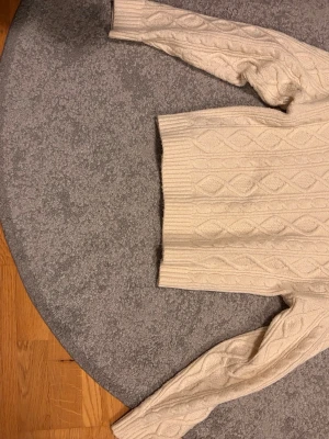 Vit kabelstickad tröja från Sthlm - Kabelstickad vit tröja från Sthlm med ribbade muddar och nederkant. Tröjan har ett klassiskt mönster med diskreta romber och är gjord i mjukt material som känns skönt mot huden. Perfekt för kyliga dagar och enkel att matcha.