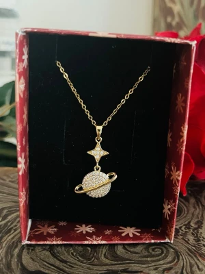 Guldigt halsband med planet-hänge - Unikt halsband i guldfärg med ett hänge som föreställer en planet med ring och en stjärna ovanför. Hänget är dekorerat med gnistrande små pärlor som ger en lyxig känsla. Perfekt accessoar för dig som gillar rymdinspirerade smycken.