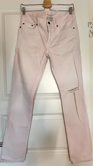 Ljusrosa jeans från Saint Laurent - Säljer ett par ljusrosa jeans från Saint Laurent Paris. Jeansen har klassisk femficksmodell, raka ben och en snygg slitning på höger lår. Tillverkade i mjuk bomull med metallknappar och bälteshällor. Perfekta för dig som vill sticka ut med en unik färg! Insömmått 72cm W29