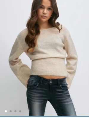 Beige stickad tröja med rund hals - En mjuk och stilren beige stickad tröja med rund hals och långa ärmar. Tröjan har en något kortare passform och är tillverkad i ett tunt, fluffigt material som ger en avslappnad look. Perfekt att matcha med jeans för en trendig outfit.