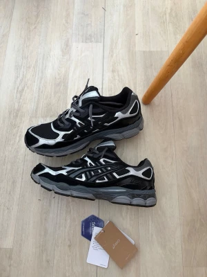 Svarta och silver Asics sneakers - Snygga Asics sneakers i svart och silver med mesh- och syntetpaneler. Skorna har en chunky sula och klassisk snörning. Ikoniska Asics-loggan syns på sidan och tungan. Perfekta för dig som gillar sportig och trendig stil.