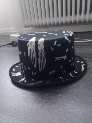 Svart cylinderhatt med skelettmotiv - Cool svart cylinderhatt med vita skelettdetaljer och benmönster tryckta över hela hatten. Perfekt accessoar till maskerad eller cosplay med halloween-tema. Hatten har en klassisk form och är tillverkad i ett glansigt tygmaterial.