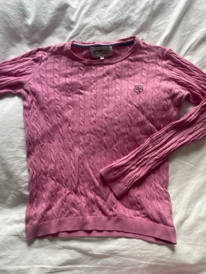 Rosa kabelstickad tröja Lexington - Säljer en rosa kabelstickad tröja från Lexington med rund halsringning och broderad logga på bröstet. Tröjan har långa ärmar och ribbade muddar vid ärmslut och nederkant. Perfekt för dig som gillar klassisk och stilren look.