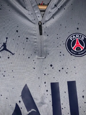 Grå PSG träningströja Jordan - Snygg grå långärmad träningströja från Jordan och Paris Saint-Germain med mörkblå logga och tryck. Tröjan har halv dragkedja, svarta stänkdetaljer och röda samt blå ränder på ärmarna. Perfekt för fotbollsträning eller gymmet.