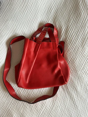 Röd handväska med nitar - Snygg röd handväska i konstläder med både axelrem och dubbla handtag. Handtagen är dekorerade med små guldfärgade nitar 