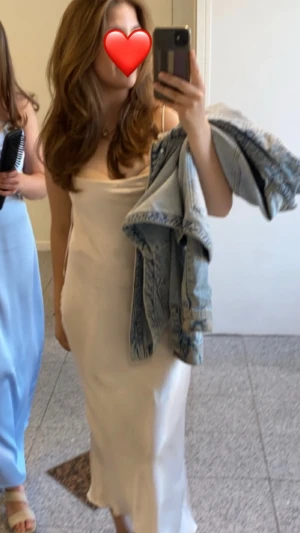 Champagnefärgad långklänning (till anklarna) med smala axelband - Elegant långklänning i champagnefärg med smala axelband och lätt draperad v-ringning. Klänningen har en slank passform och är tillverkad i ett glansigt satinliknande material som ger en lyxig känsla. Perfekt för dig som vill ha en stilren och modern look.