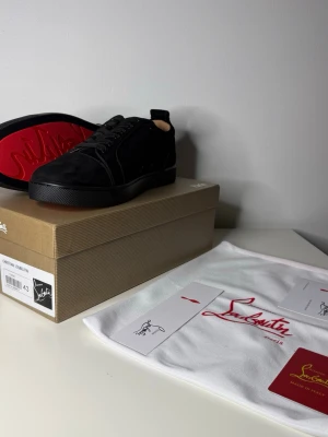 Svarta sneakers från Christian Louboutin - Stilrena svarta sneakers från Christian Louboutin med klassisk röd sula och diskret logga. Skorna har en låg profil, svarta skosnören och ovandel i mocka. Perfekta för dig som vill ha en lyxig och tidlös sneaker med ikonisk design.