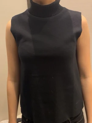 Svart ärmlös polotopp - Snygg svart polotopp med ribbad hög krage och ärmlös design. Perfekt att styla med jeans eller kjol för en clean och modern look. Materialet känns mjukt och stretchigt.