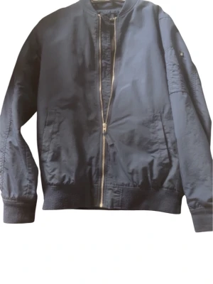 Mörkblå bomberjacka med dragkedja - Snygg mörkblå bomberjacka med klassisk passform och guldfärgad dragkedja framtill. Jackan har ribbade muddar vid ärmslut och nederkant samt fickor på sidorna. Materialet är slätt och jackan har en cool, urban look.