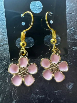 Rosa blomörhängen med gulddetaljer - Snygga örhängen i form av blommor med fem rosa kronblad och guldfärgad kant. Blommornas mitt har små guldfärgade detaljer som ger extra glans. Krokarna är också guldfärgade och ger en trendig look. Perfekta för dig som vill ha en söt och färgglad accessoar.