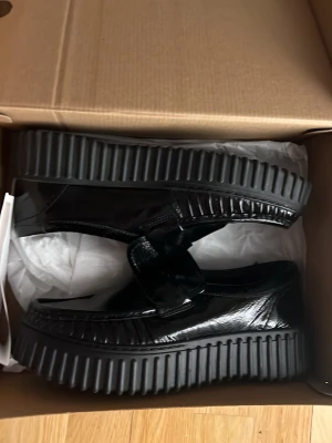 Svarta chunky loafers i lack - Säljer ett par svarta loafers med chunky sula och lackad finish. Skorna har en bred rem över foten. Materialet är glansigt och sulan är rejält räfflad. Köpte för 1399 kr men tyvärr behöver dom säljas för att storleken inte passar. Sticket är helt nytt, använt bara ett fåtal gånger.