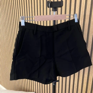 Svarta kostymshorts från H&M - Stilrena svarta shorts med kostymkänsla, pressveck framtill och sidofickor. De har hällor för bälte och två dekorativa bakfickor. Perfekta för en dressad look och passar till många olika outfits.