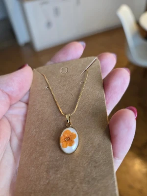 Guldigt halsband med orange blomma - Snyggt halsband med tunn guldfärgad kedja och ett ovalt hänge i vitt med en orange blomma. Perfekt accessoar för dig som gillar unika smycken med blommotiv. Materiall-rostfritt stål, haldkedja 40+ 5 cm