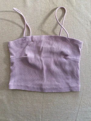 Ljuslila ribbad linnetopp - Säljer en ljuslila ribbad linnetopp med smala axelband i storlek XS. Toppen är croppad och har en enkel, stilren design som passar perfekt till sommaren. Materialet är mjukt och stretchigt för en bekväm passform.