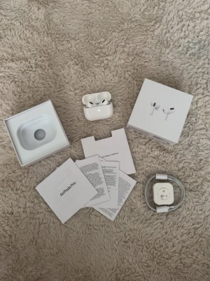 Apple AirPods Pro med laddningsetui - Säljer ett par Apple AirPods Pro i vit färg med tillhörande trådlöst laddningsetui. Hörlurarna har en stilren och kompakt design med silikonproppar och levereras med originalkartong, laddkabel och manualer. Perfekta för musik, samtal och träning.