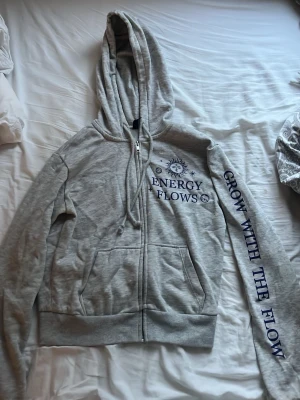 Grå hoodie med tryck och dragkedja - Säljer en grå hoodie med dragkedja och huva. På bröstet står det 'ENERGY FLOWS' med sol- och mån-symboler, och på ena ärmen står det 'GROW WITH THE FLOW' i blå text. Hoodien har fickor framtill och justerbara snören i huvan. Perfekt för chill dagar.