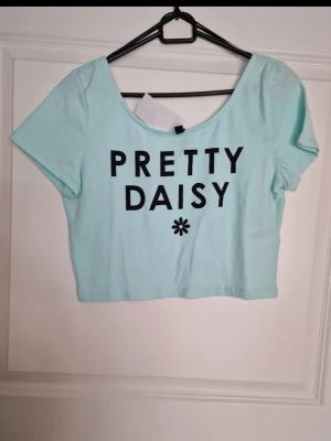 Ny mint croppad topp Pretty Daisy med etikett - Söt ljusblå croppad topp med svart texten 'PRETTY DAISY' och en liten blomdetalj framtill. Toppen har rund halsringning och korta ärmar. Perfekt för dig som gillar en enkel men trendig stil.