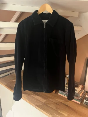 Svart manchester overshirt med dragkedja - Stilren mörkblå overshirt i manchestertyg med dragkedja framtill och klassisk krage från Bläck. Bröstficka på vänster sida och knappar vid ärmsluten ger en clean look. Perfekt lager-på-lager-plagg för dig som gillar minimalistisk stil. Jackan är st Small men passar som en Medium. Passar perfekt för de varmare dagarna som är på väg. Skicket på jackan ör inprincip ny har använt väldigt lite. Hör av er vid mer frågor😀