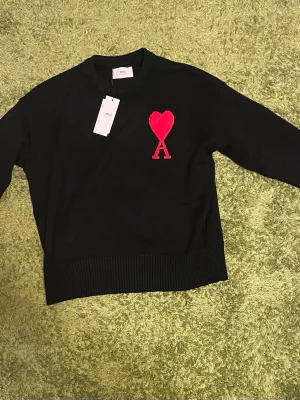 Svart stickad tröja från Ami Paris - Svart stickad tröja från Ami Paris med röd hjärtlogga och bokstaven A broderad på bröstet. Klassisk rund halsringning och ribbade muddar. Perfekt för dig som gillar stilrena plagg med en twist.