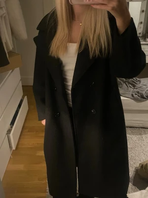 Svart lång kappa med dubbelknäppning - Stilren svart kappa med dubbelknäppning och bred krage. Lång modell som ger en snygg och klassisk siluett. Perfekt för dig som gillar minimalistisk och tidlös stil. Materialet ser ut att vara ullblandning.