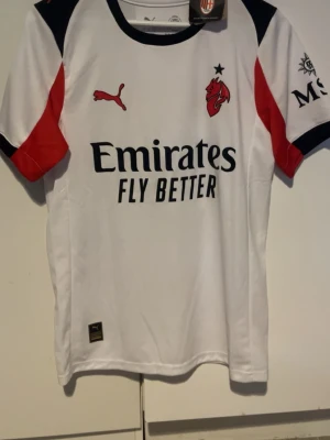 AC Milan bortatröja Puma Rafa Leão - Snygg vit AC Milan bortatröja från Puma med svarta och röda detaljer. Tryck med Emirates Fly Better på framsidan och Rafa Leão 10 på ryggen. Korta ärmar, klubbmärke och Puma-logga på bröstet. Perfekt för fotbollsfans!