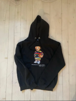 Svart hoodie Polo Bear Ralph Lauren - Svart hoodie från Ralph Lauren med den ikoniska Polo Bear-trycket på bröstet. Björnen har färgglada kläder och håller en basketboll. Hoodien har dragsko i huvan och ribbade muddar. Perfekt för dig som gillar streetwear och vill sticka ut. Står att de är M men passar bättre folk som har S 