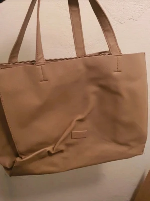 Beige shoppingväska - Stilren beige shoppingväska i skinnimitation. Väskan har dubbla handtag, dragkedja upptill och en enkel, rektangulär form. Perfekt för att bära med sig allt man behöver under dagen.