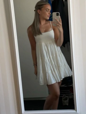 Vit kort sommarklänning med volang Zara - Säljer en vit kort klänning med smock upptill och tunna axelband. Klänningen har volangdetaljer och är perfekt för varma dagar. Den är luftig och har en avslappnad passform.