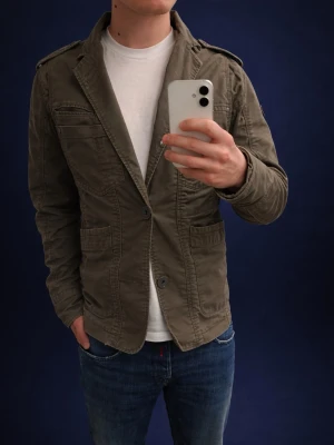 Olivgrön blazerjacka - Snygg olivgrön blazerjacka från Jack & Jones med klassisk krage, axelklaffar och flera fickor framtill. Jackan har knappstängning och är tillverkad i bomull för en bekväm känsla. Perfekt för en avslappnad men stilren look.