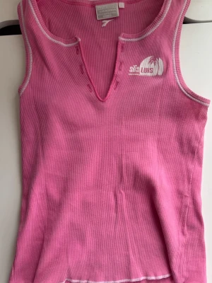Rosa ribbad linnetopp - Säljer en rosa ribbad linnetopp med vit logga på bröstet och dekorativa knappar vid den v-ringade halsen. Toppen är ärmlös och har vita kontrastsömmar längs kanterna. Perfekt för en avslappnad och trendig look.