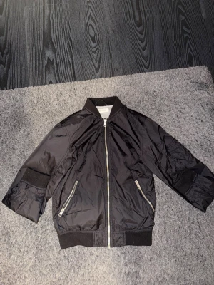 Svart bomberjacka från H&M - Snygg svart bomberjacka från H&M med silvriga dragkedjor och ribbade muddar vid ärmslut och nederkant. Jackan har två fickor med dragkedja och klassisk bomberkrage. Materialet är glansigt och lätt, perfekt för en cool streetstil.