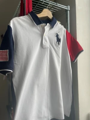 Pikétröja från Polo Ralph Lauren - Snygg pikétröja från Polo Ralph Lauren med vit framsida, mörkblå rygg och ena ärmen röd. Stor logga på bröstet, amerikansk flagga på ärmen och stor siffra 3 på ryggen. Klassisk krage och knappar framtill. Perfekt för en sportig och stilren look.