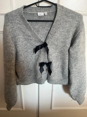 Grå kofta med rosetter från Name It - Söt grå kofta från Name It med två svarta rosetter framtill. Koftan har v-ringning, långa ärmar och ribbade muddar. Perfekt för lager på lager och enkel att matcha med olika outfits.