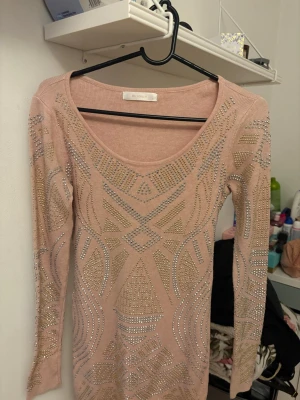 Rosa topp med guld- och silverdetaljer - Trendig rosa topp från Mc Lorene med långa ärmar och snyggt mönster av guld- och silverfärgade nitar. Toppen har en djup rundringning och är stretchig för en tight passform. Perfekt för dig som gillar att sticka ut med coola detaljer.