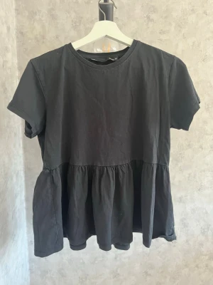 Svart volangblus - Svart blus med rund hals och korta ärmar. Toppen har en lös passform och volang nedtill som ger en söt och avslappnad look. Kläderna stryks och noppras alltid innan köp🎀