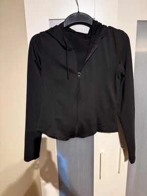 Svart croppad hoodie med dragkedja - Snygg svart croppad hoodie med dragkedja framtill och långa ärmar. Hoodien har en klassisk huva med dragsko och är tillverkad i ett mjukt, stretchigt material som känns bekvämt mot huden. Perfekt för en avslappnad och trendig look.