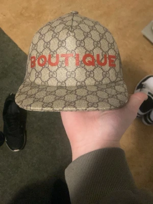Gucci keps med BOUTIQUE-tryck - Säljer en beige keps från Gucci med det klassiska GG-mönstret över hela kepsen och röd text 'BOUTIQUE' framtill. Kepsen har böjd skärm och justerbar rem bak. Materialet är canvas med exklusiv känsla och streetwear-vibe.