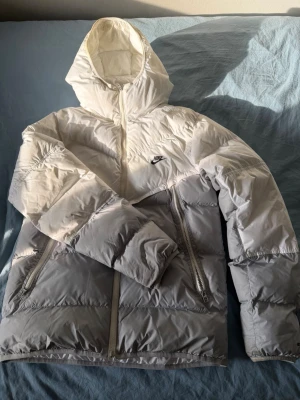 Nike tvåfärgad pufferjacka med huva - Säljer en riktigt snygg pufferjacka från Nike i vitt och ljusgrått. Jackan har en stor huva, två dragkedjeförsedda fickor framtill och Nike-logga på bröstet. Perfekt för kalla dagar när du vill hålla stilen och värmen. Jackan är köpt för 2100 kr, och mitt pris är 700 kr. Använt bara 1 vinter och är väl skött.